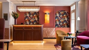A reception area with a wooden desk, a chandelier, and floral wall art. - Mayflower Opéra  (Paris)