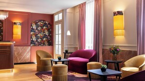 A hotel lobby with a wooden reception desk, plush seating, a large floral wall mural, and a large window with curtains. - Mayflower Opéra  (Paris)