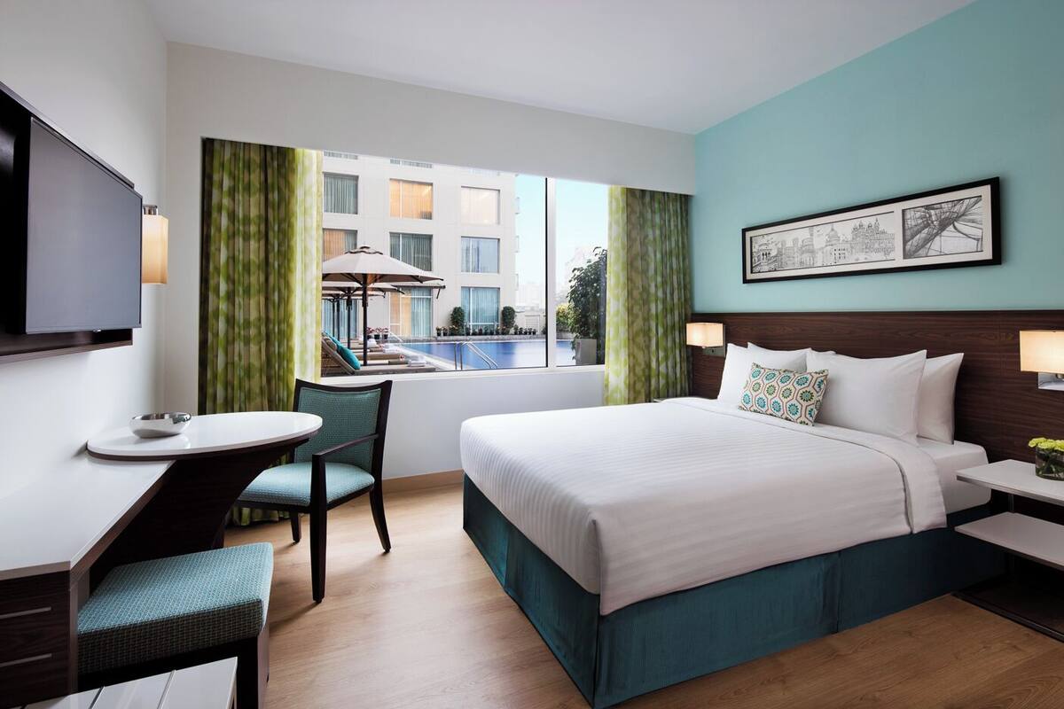 Habitación de hotel con cama, escritorio, silla, televisor, ventana con vista a la piscina y un cuadro en la pared.