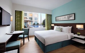 Habitación de hotel con cama, escritorio, silla, televisor, ventana con vista a la piscina y un cuadro en la pared.
