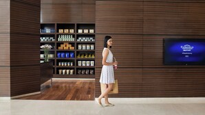Een vrouw loopt door een moderne hotellobby met een groot televisiescherm waarop een boodschap staat, houten lambrisering en een netjes ingerichte plank met diverse spullen.