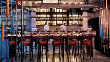 Un bar moderno con mesas de madera y sillas con cojines rojos, un ambiente bien iluminado y un cantinero preparando bebidas.