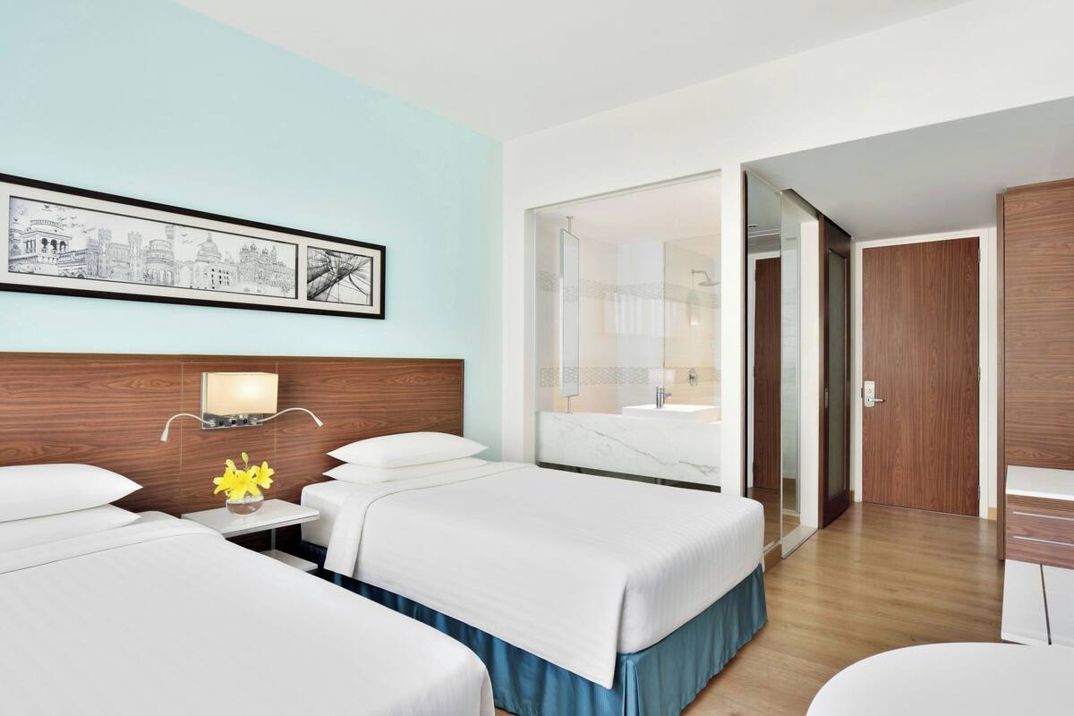 Habitación de hotel con dos camas, una mesita de noche con lámpara, una obra de arte en la pared y un baño visible a través de una puerta de cristal.