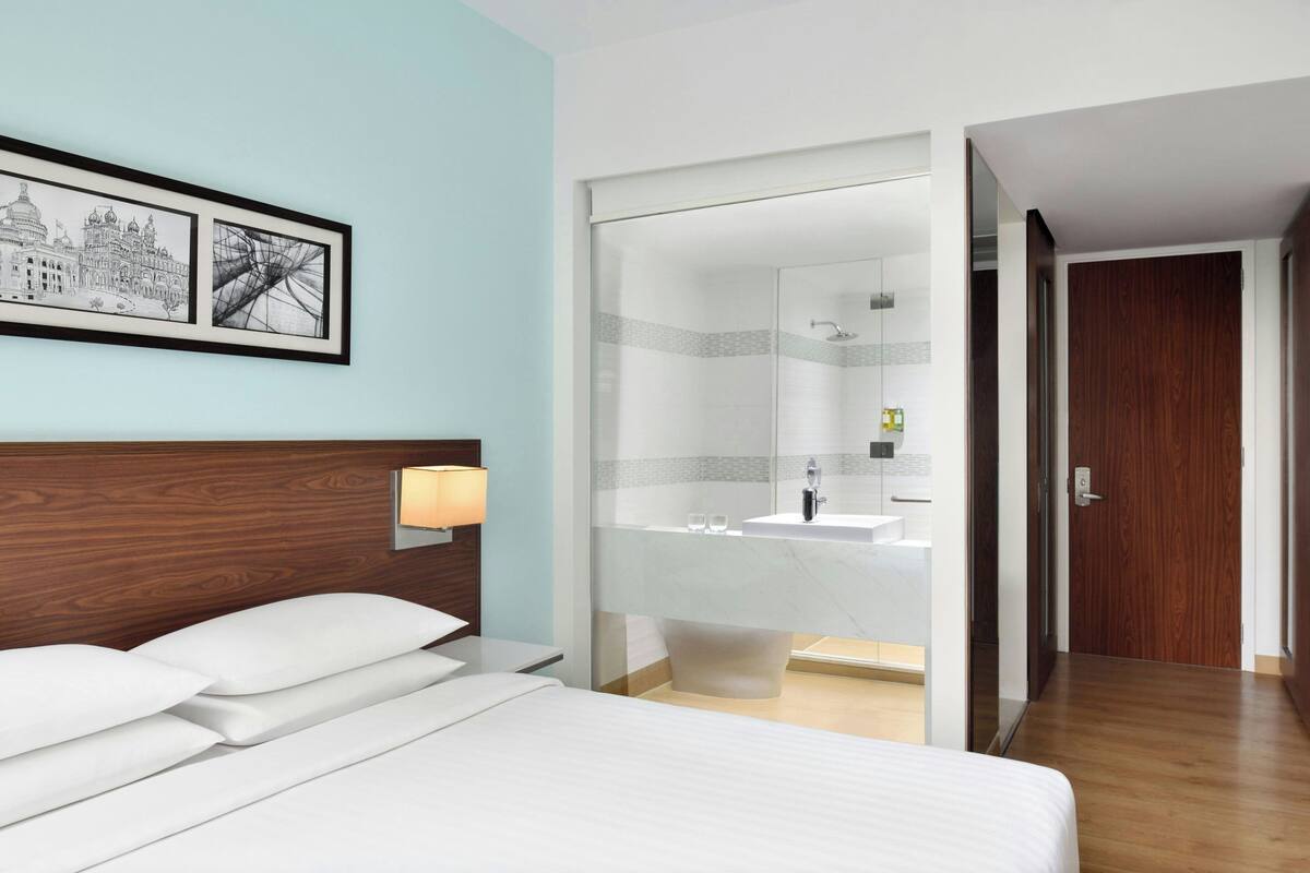 Habitación de hotel con cama, mesita de noche con lámpara, una obra de arte urbana enmarco, baño con separador de vidrio y puerta de madera.