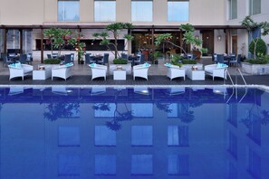 Un área junto a la piscina con sillones de descanso y mesas, una piscina cristalina y un edificio con grandes ventanales al fondo.