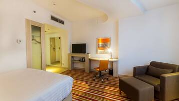 Quarto de hotel com cama, escrivaninha com cadeira, televisão e sofá.