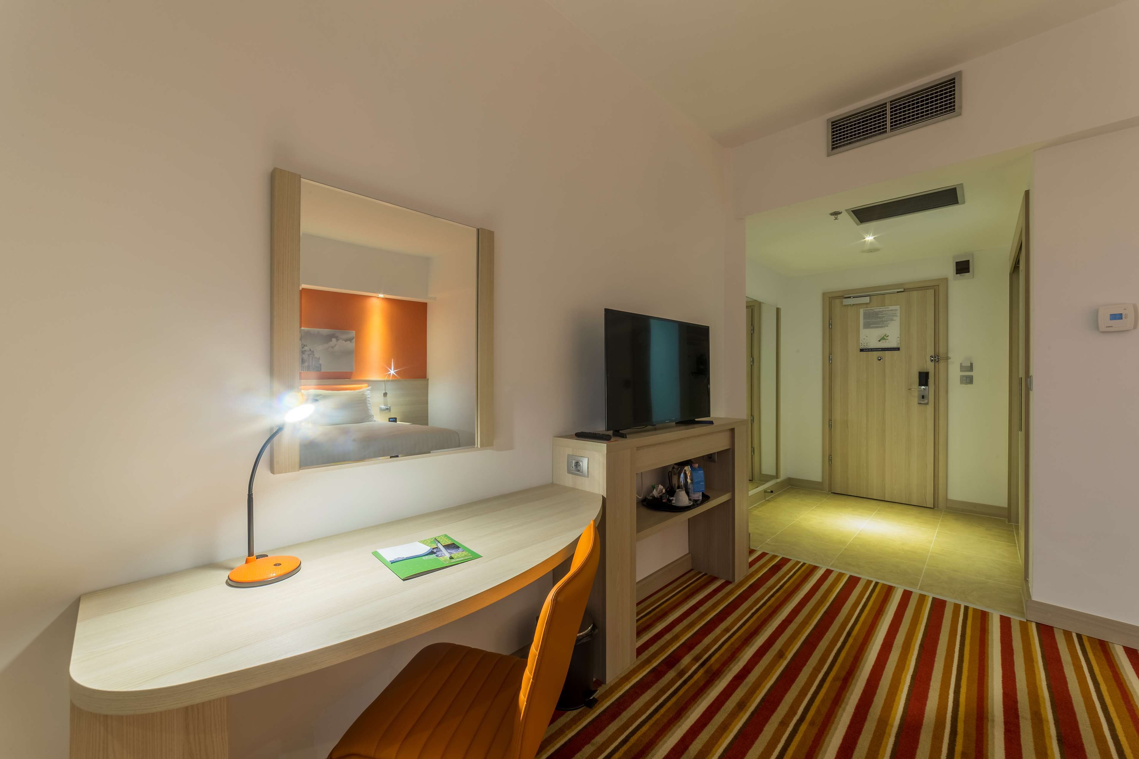 A hotel room with a desk, chair, television, and a mirror.