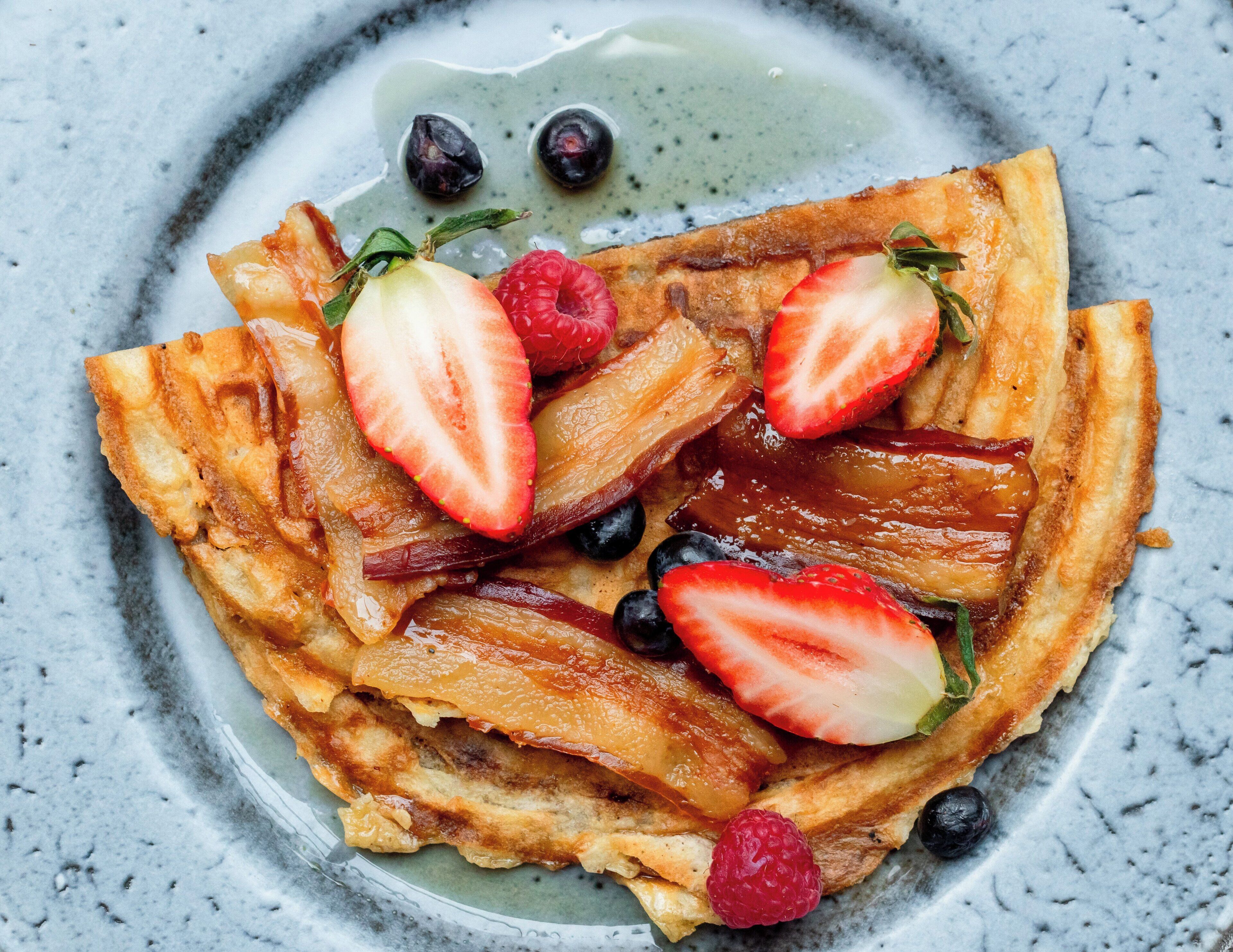 A plate of pancakes topped with strawberries, raspberries, and blueberries, with syrup on a marble surface.