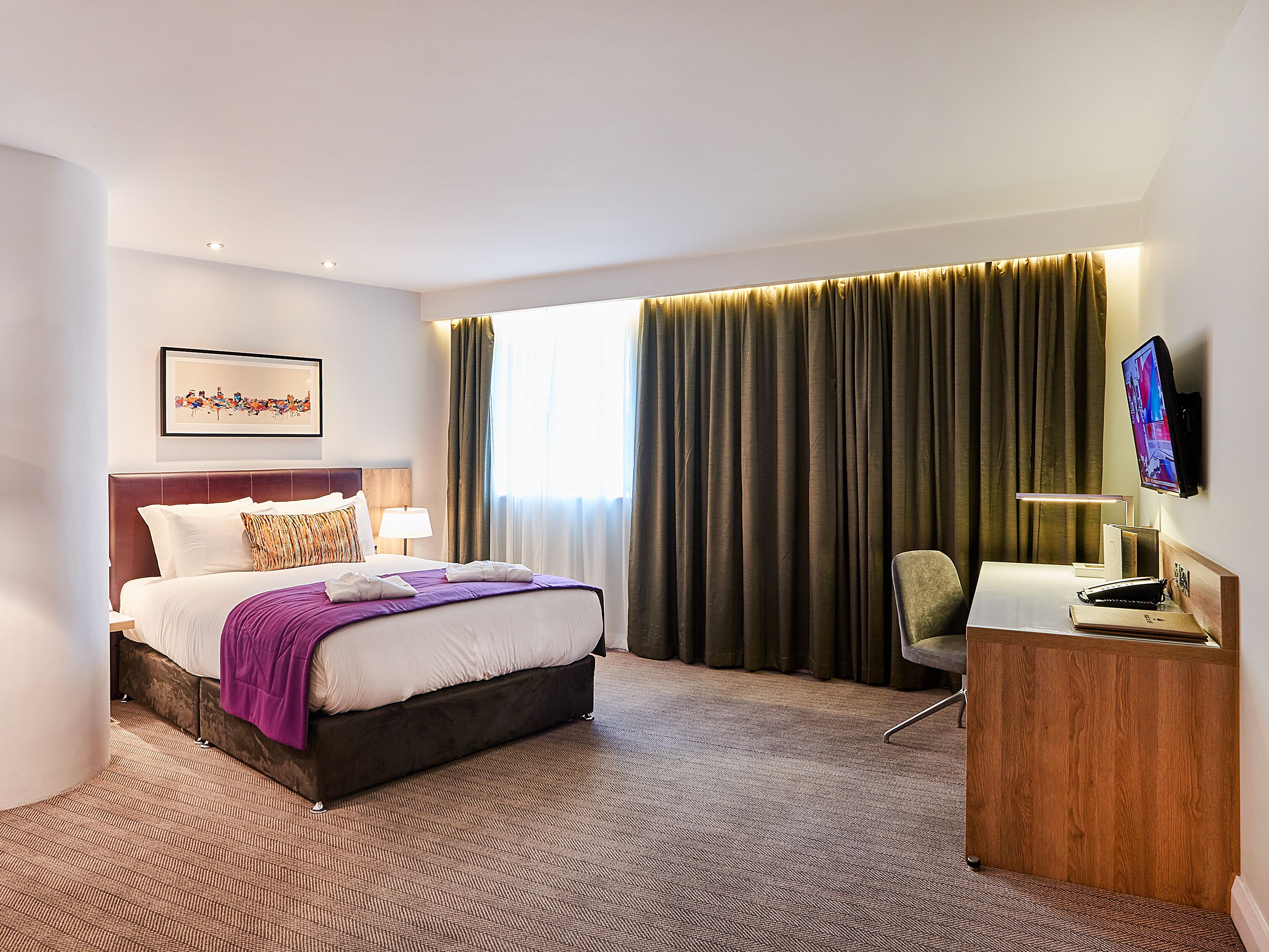 A hotel room with a bed, a desk with a lamp, a TV, and a window with curtains.