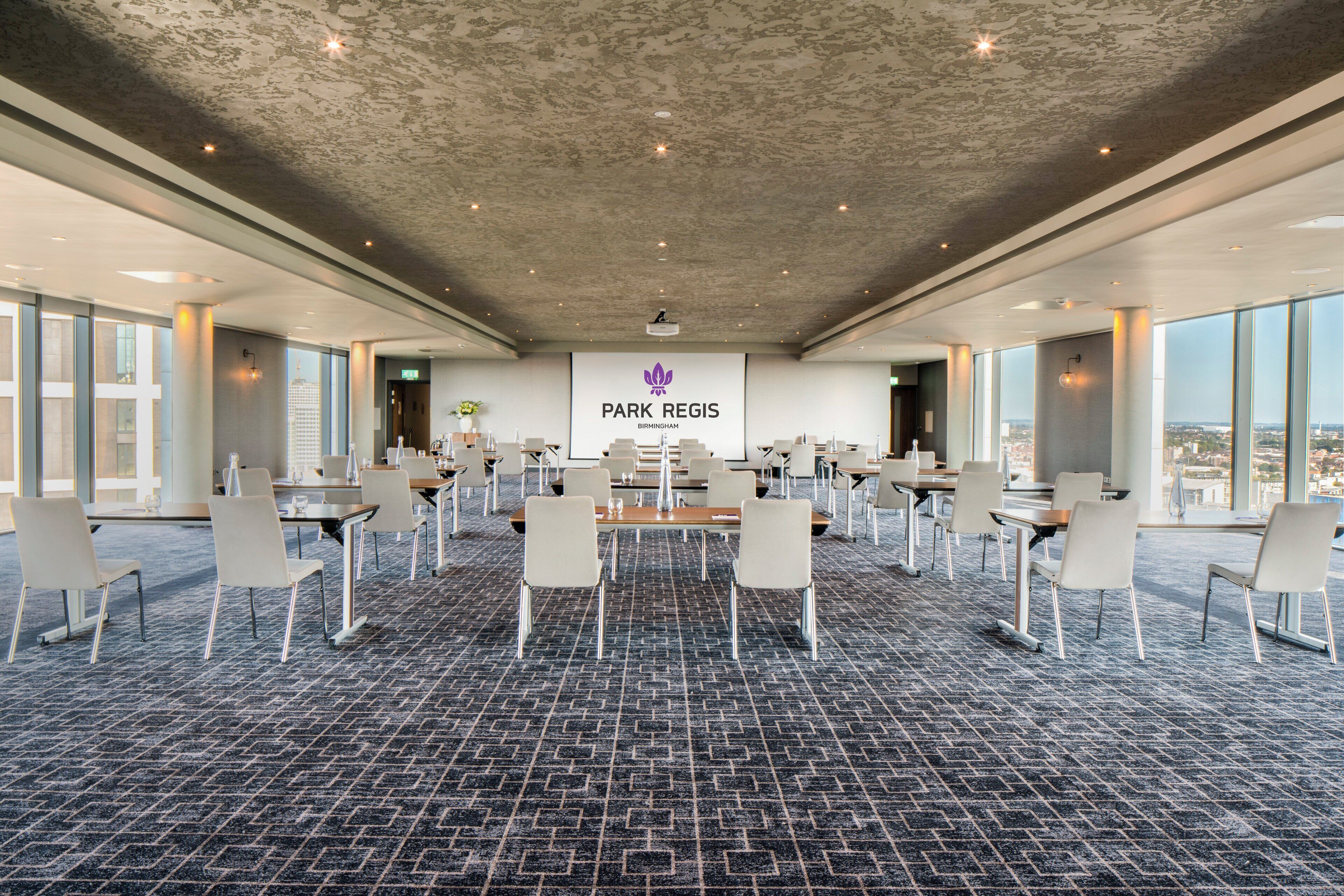 A conference room with a carpeted floor, white chairs, and tables. A banner with "Park Regis Birmingham" is visible.