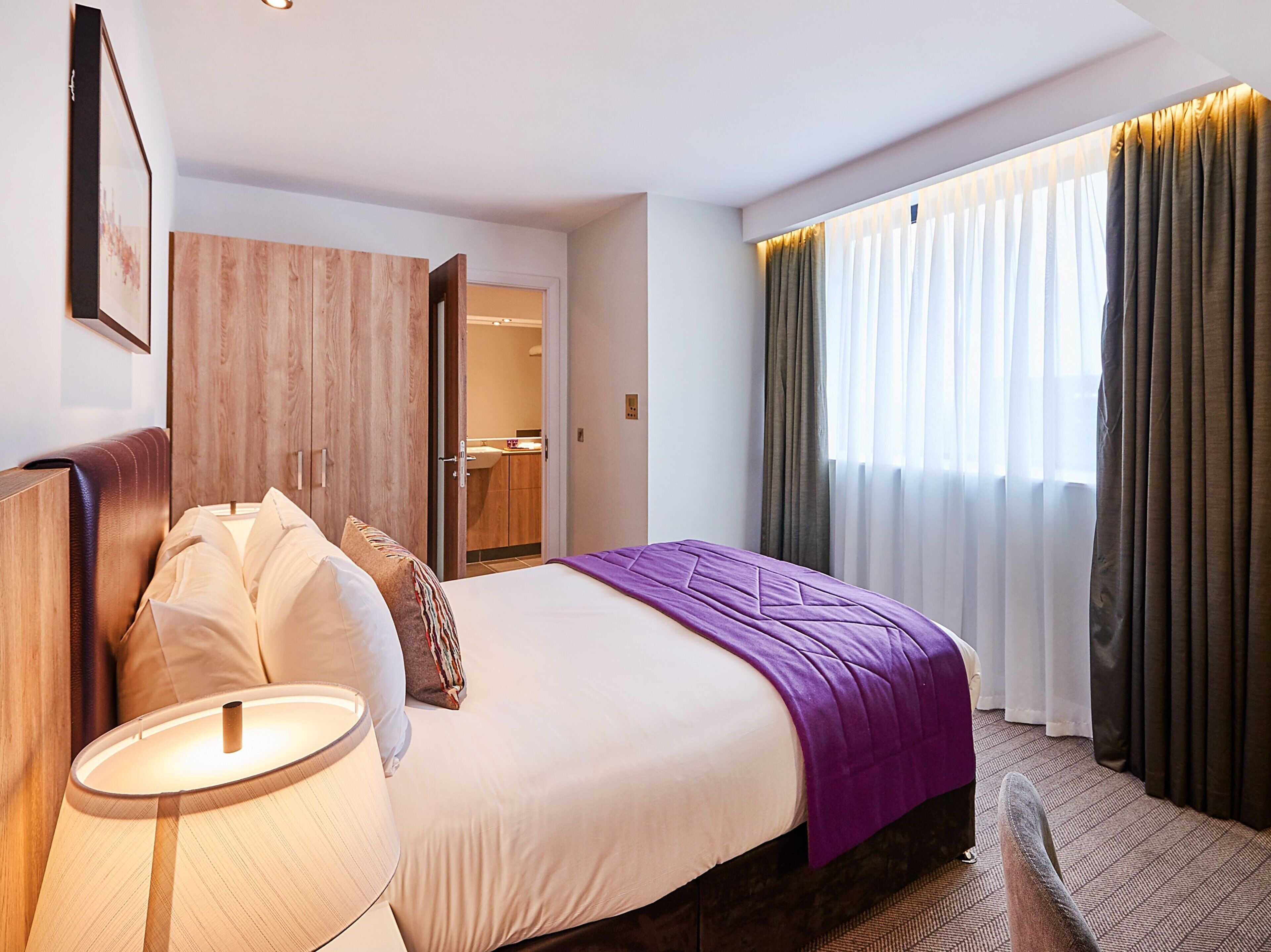 A hotel room with a bed, a bedside lamp, a wooden wardrobe, and a window with curtains.