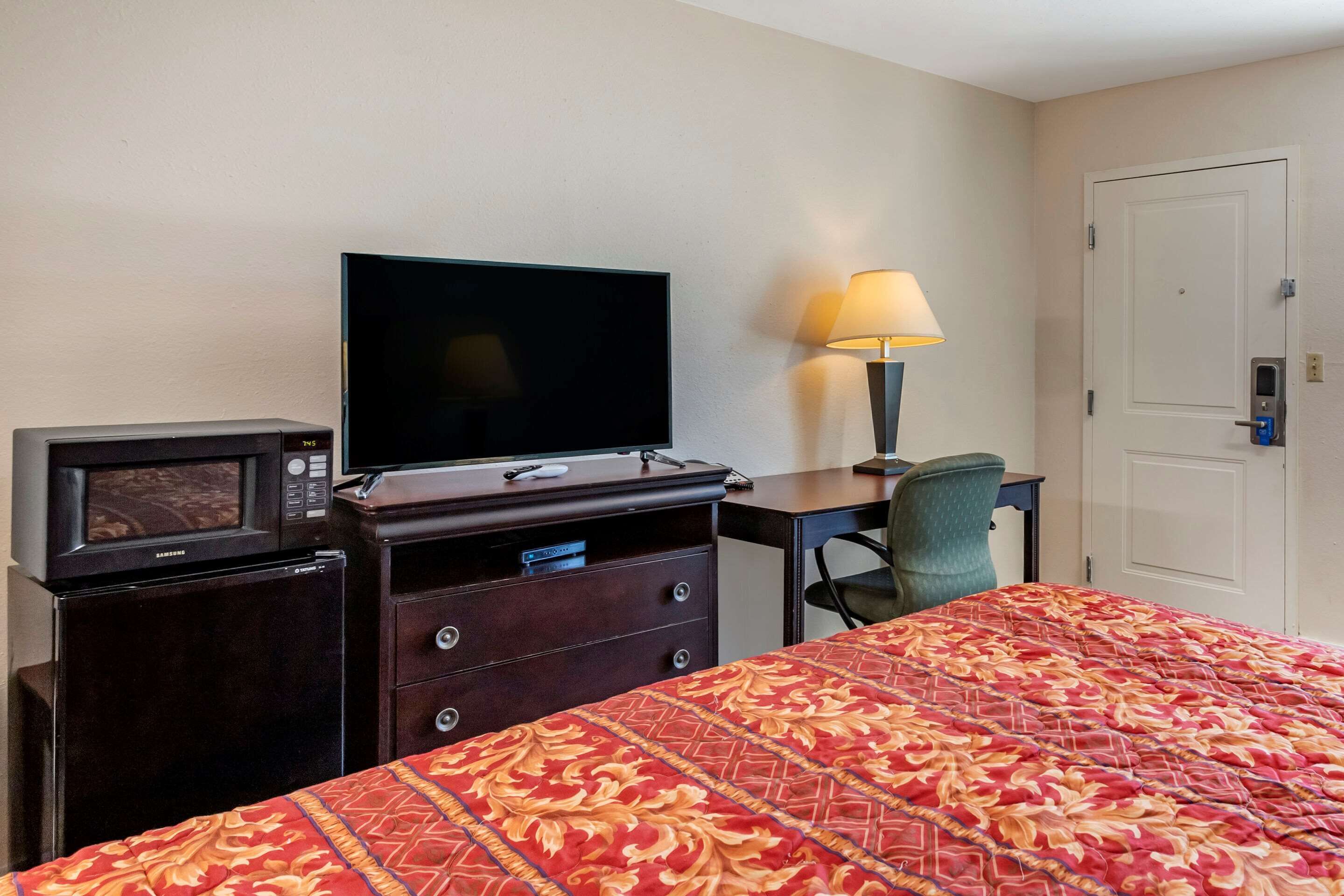 A hotel room with a bed, a TV, a microwave, a desk with a lamp, and a door.