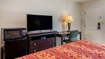 A hotel room with a bed, a TV, a microwave, a desk with a lamp, and a door.