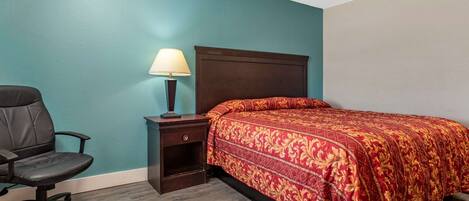 Un dormitorio con paredes azul verdosas, cabecera de madera, colcha estampada roja, una silla de escritorio negra y una mesita de noche con lámpara.