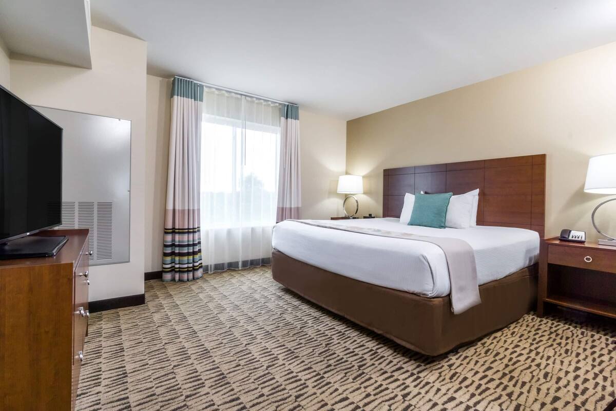 Ein Hotelzimmer mit Bett, Schreibtisch, Fernseher, Lampe und einem fenster mit Vorhängen.