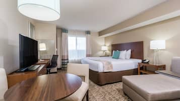 A hotel room with a bed, a desk with a TV, a chair, a table, and a lamp.