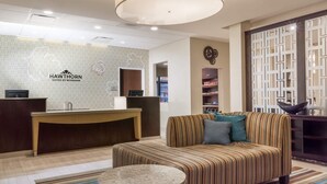 Zona de recepción de un hotel con mostrador, un sofá, una mesita redonda y una pared decorativa con un logotipo.