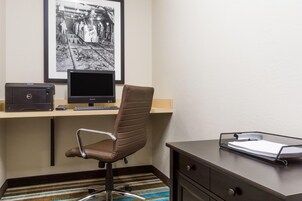 A hotel room with a desk, chair, computer, printer, and a framed photo on the wall.