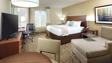 A hotel room with a large bed, a desk with a computer, a TV, a chair, a sofa, and a painting on the wall.