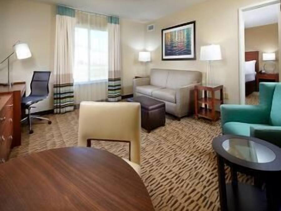 A hotel room with a round wooden table, a sofa, a chair, a small ottoman, a glass coffee table, and a lamp.