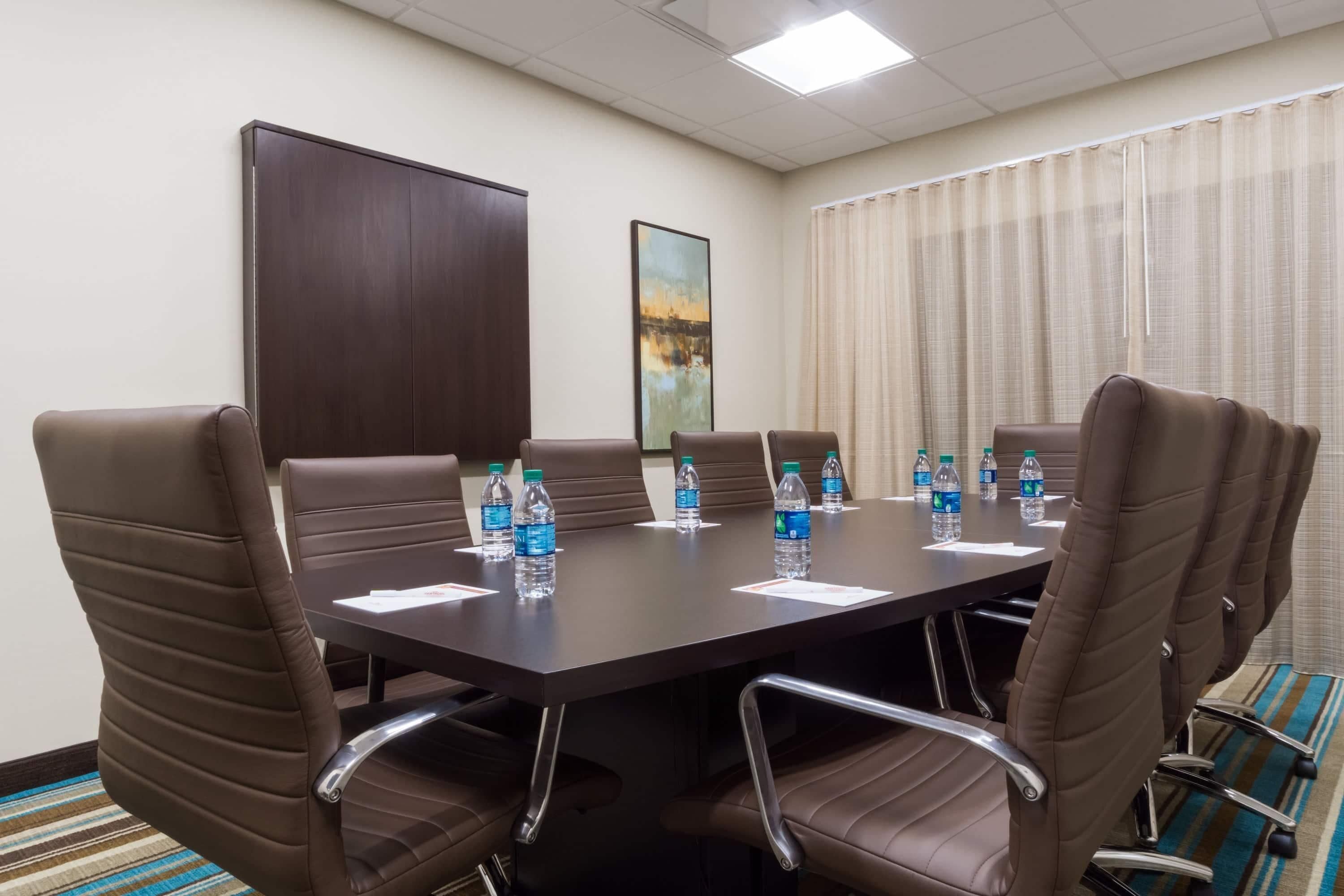 Una sala de conferencias con una mesa de madera oscura, sillas de cuero marrón, botellas de agua y un cuadro en la pared.