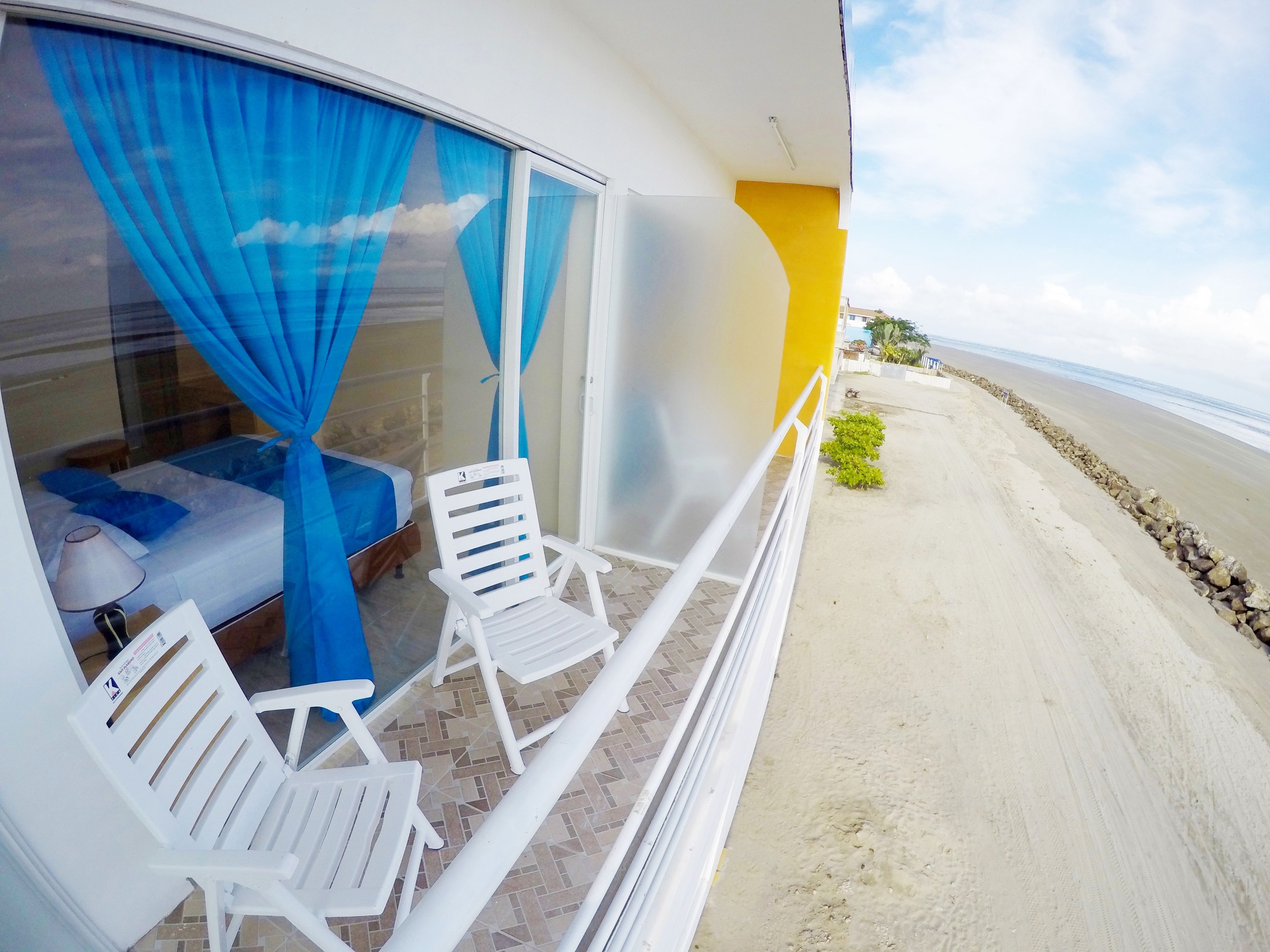 A balcony with white chairs, a bed with blue curtains, and a view of the beach.