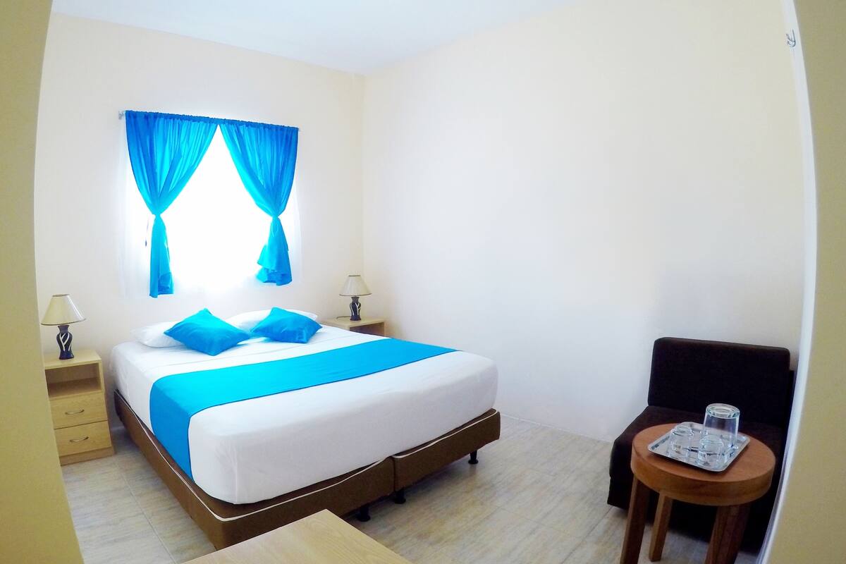 A hotel room with a bed, two bedside tables with lamps, a small round table with glasses, and a window with blue curtains.