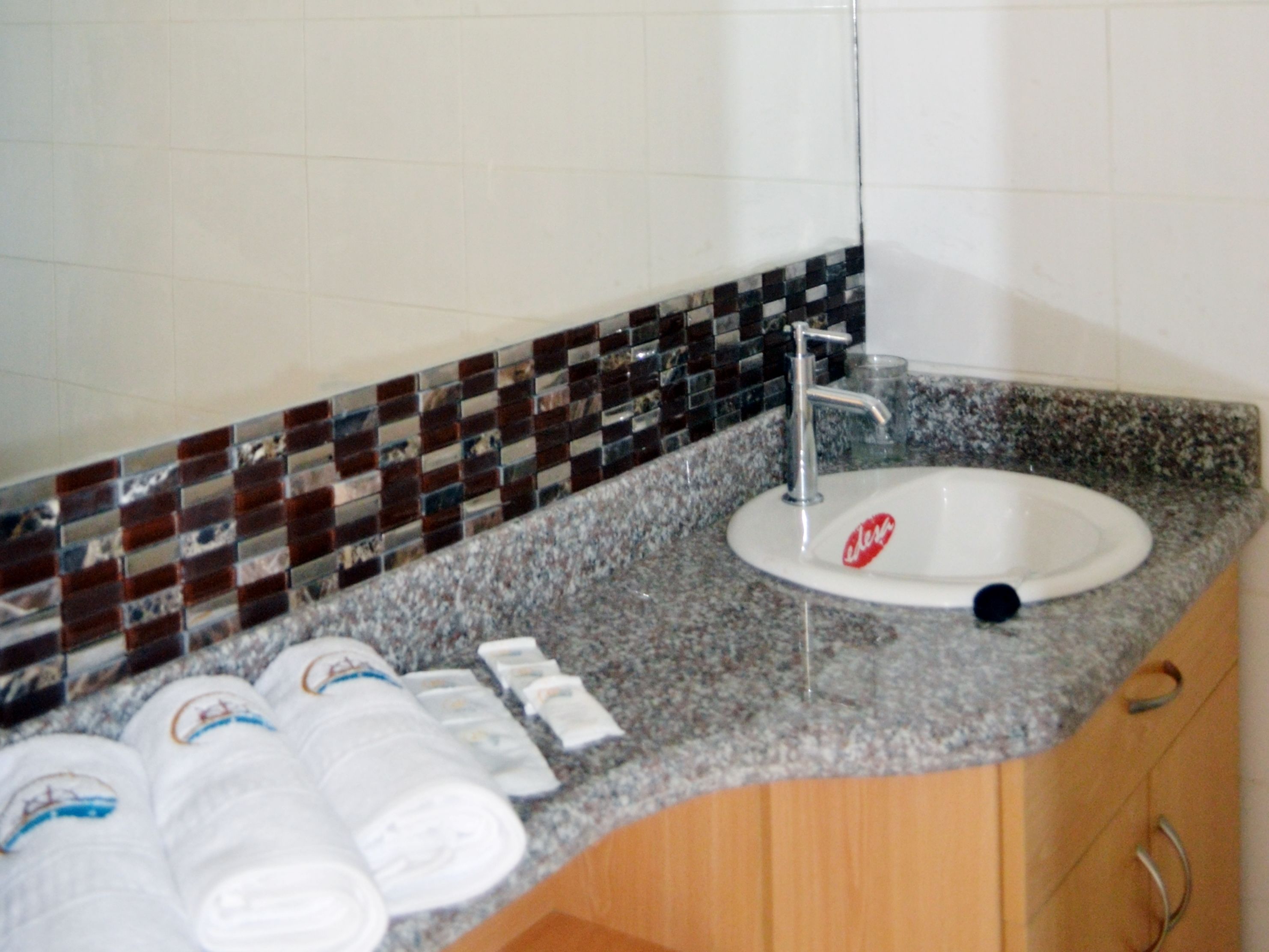 Casa de banho com bancada de granito, lavatório branco e painel de azulejos mosaicos.