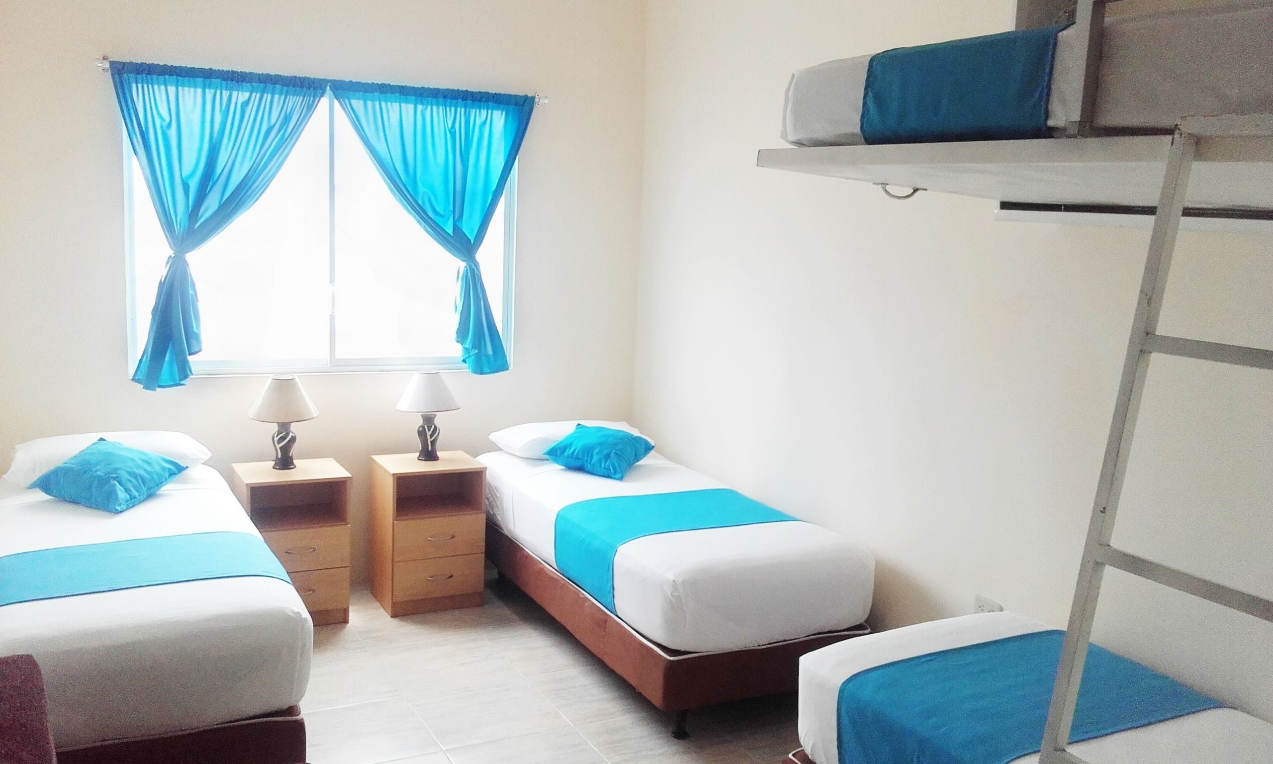 Una habitació amb dos llits individuals, cadascun amb un llençol blau i una almoçada, una finestra amb cortines blaves, i una lliterà sobre la dreta.