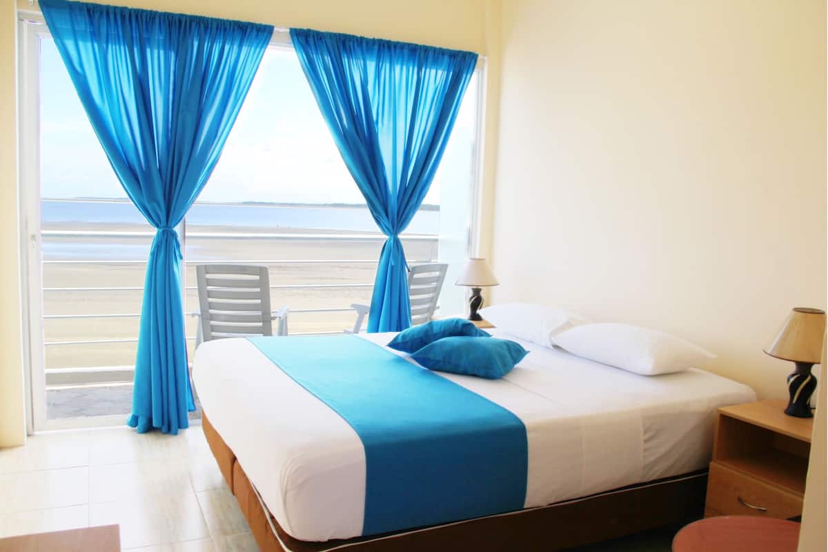 A hotel room with a bed, a nightstand, a lamp, a chair, and a view of the ocean through large windows with blue curtains.