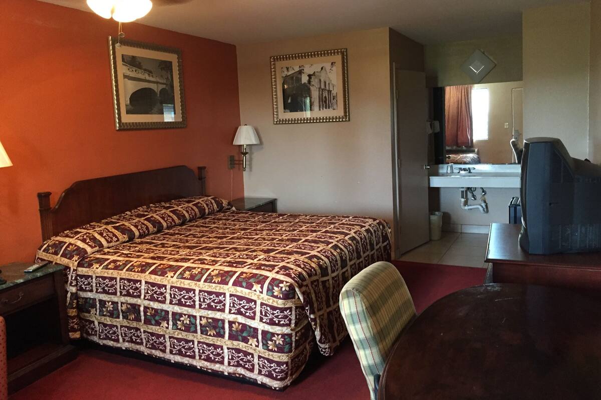Hotelkamer met een groot bed, een bureau, een stoel, een plafondventilator en een badkamer met een wastafel en spiegel.