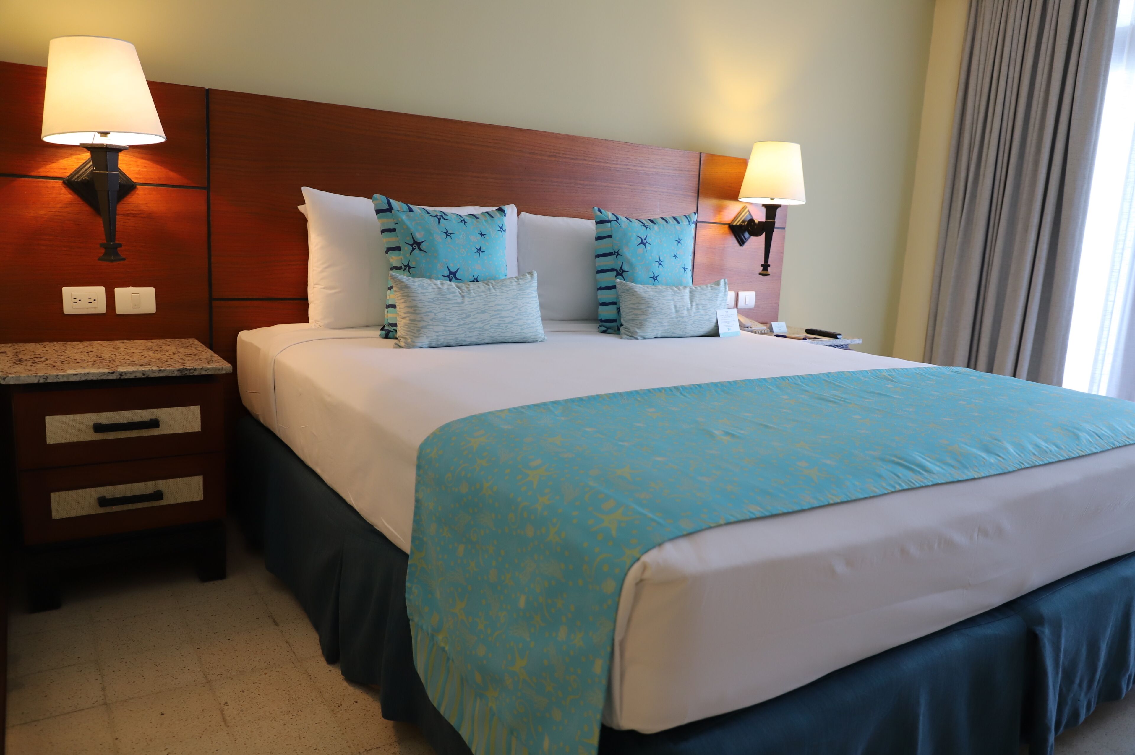 A hotel room with a large bed, two bedside tables, two wall-mounted lamps, and a window with curtains.