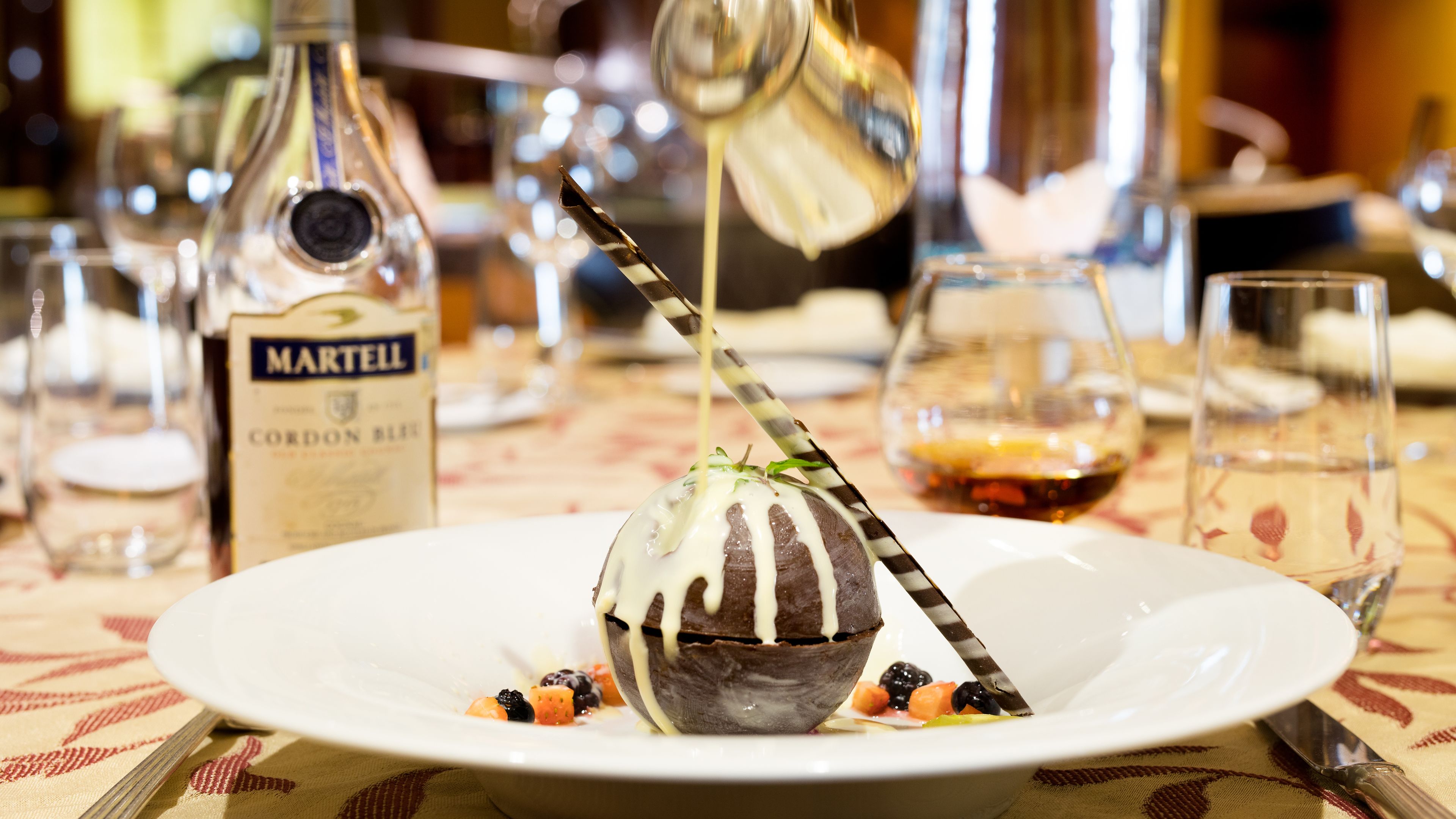Un postre sobre un plato blanco con una cuchara a rayas, rodeado de varias botellas y vasos sobre un mantel.