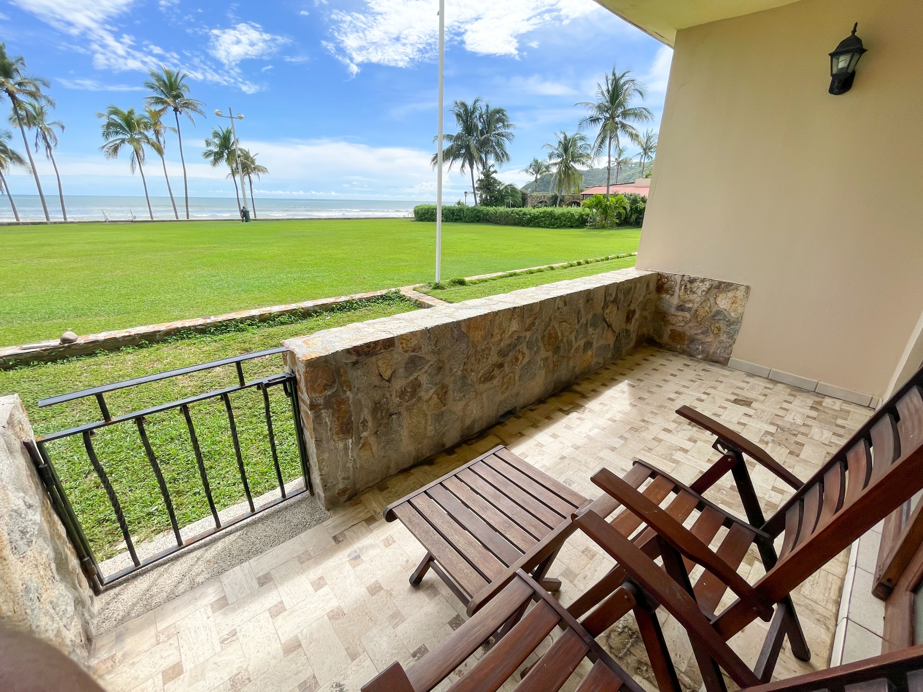 A balcony with a stone wall, wooden furniture, and a view of the ocean and palm trees.