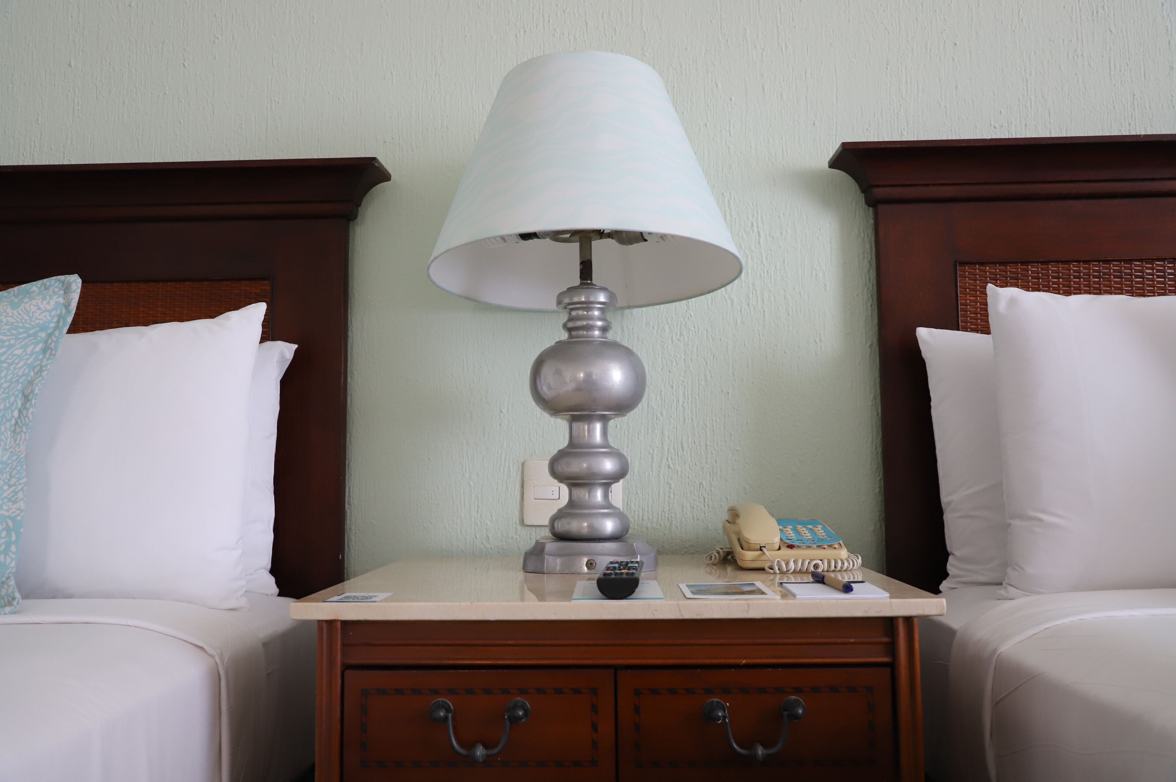 Habitación de hotel con cama, mesita de noche con lámpara, teléfono y una pequeña mesa con control remoto.