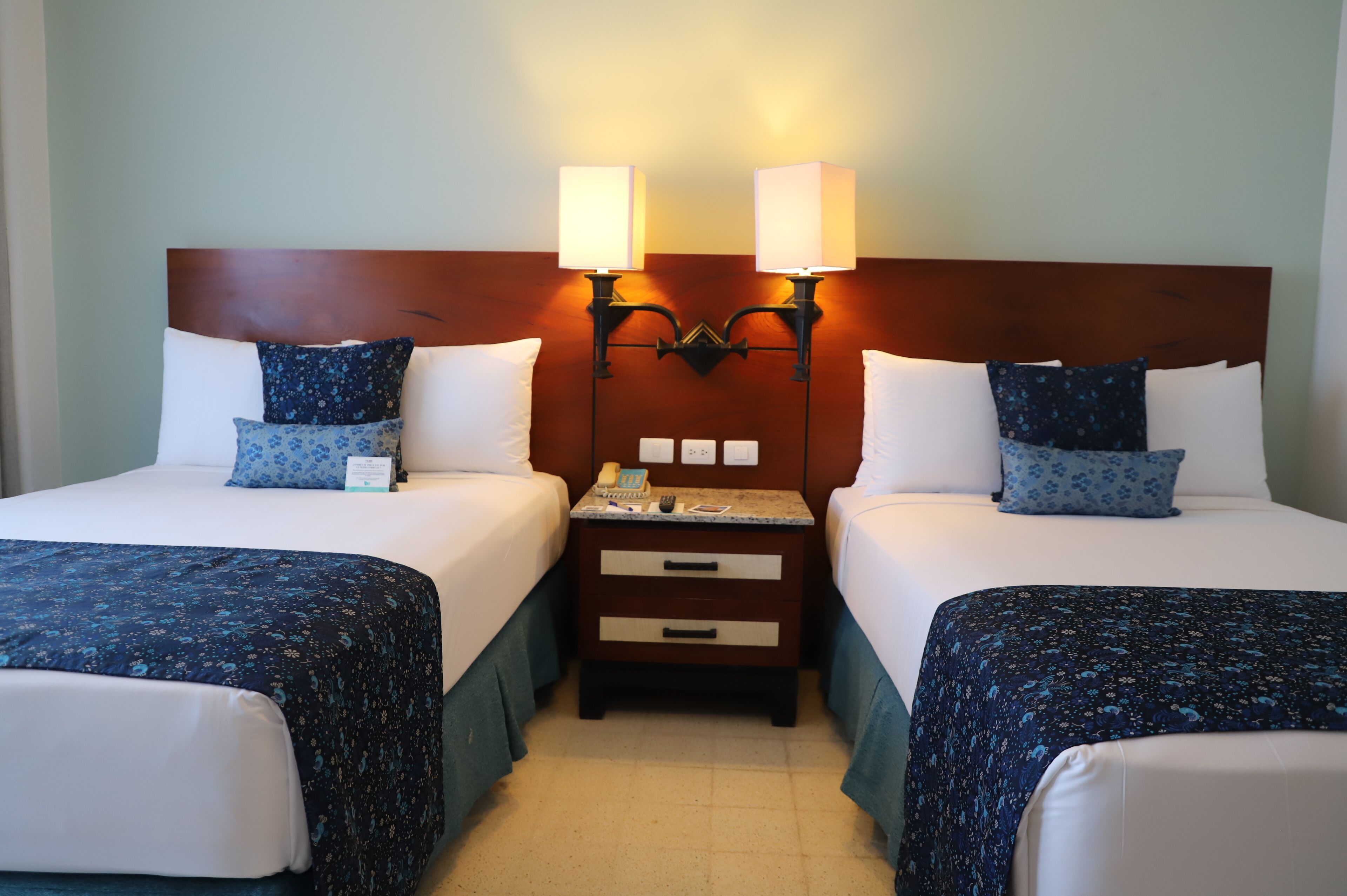 A hotel room with two beds, each with a floral-patterned bedspread, a wooden headboard, bedside lamps, and a small bedside table with a phone and a book.