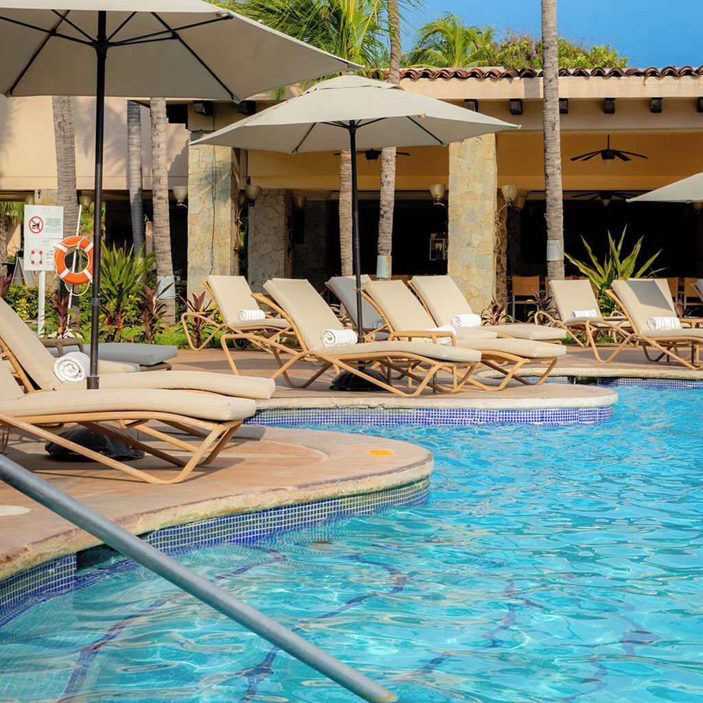 Zona de piscina con sillones de descanso y sombrillas, rodeada de palmeras y un edificio con techo de tejas.