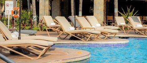 Zona de piscina con sillones de descanso y sombrillas, rodeada de palmeras y un edificio con techo de tejas.