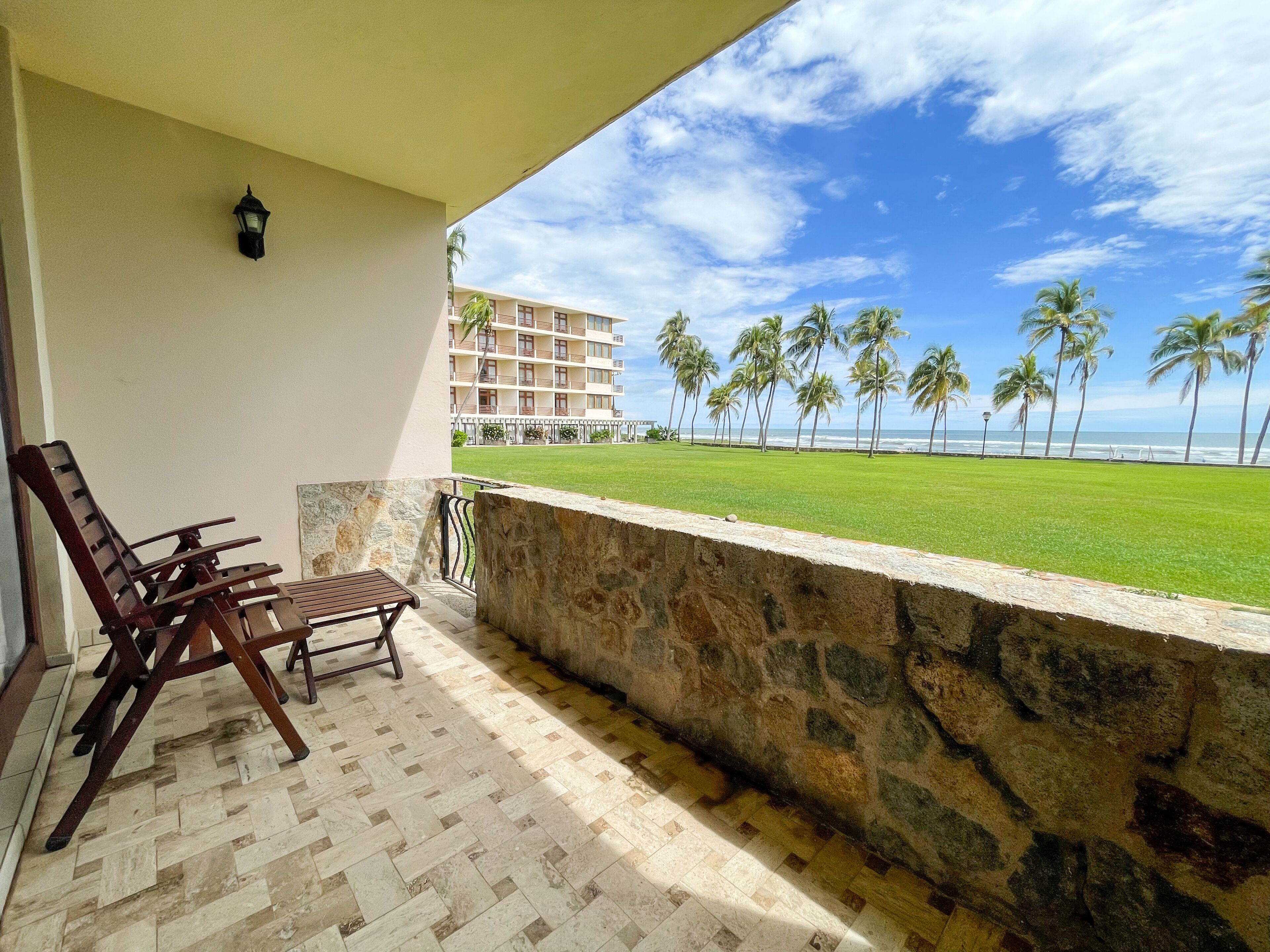 A balcony with a stone wall, wooden chairs, and a view of a beach with palm trees and a building in the background.