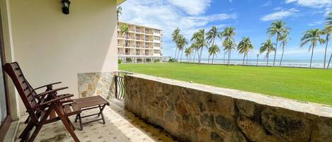 A balcony with a stone wall, wooden chairs, and a view of a beach with palm trees and a building in the background.