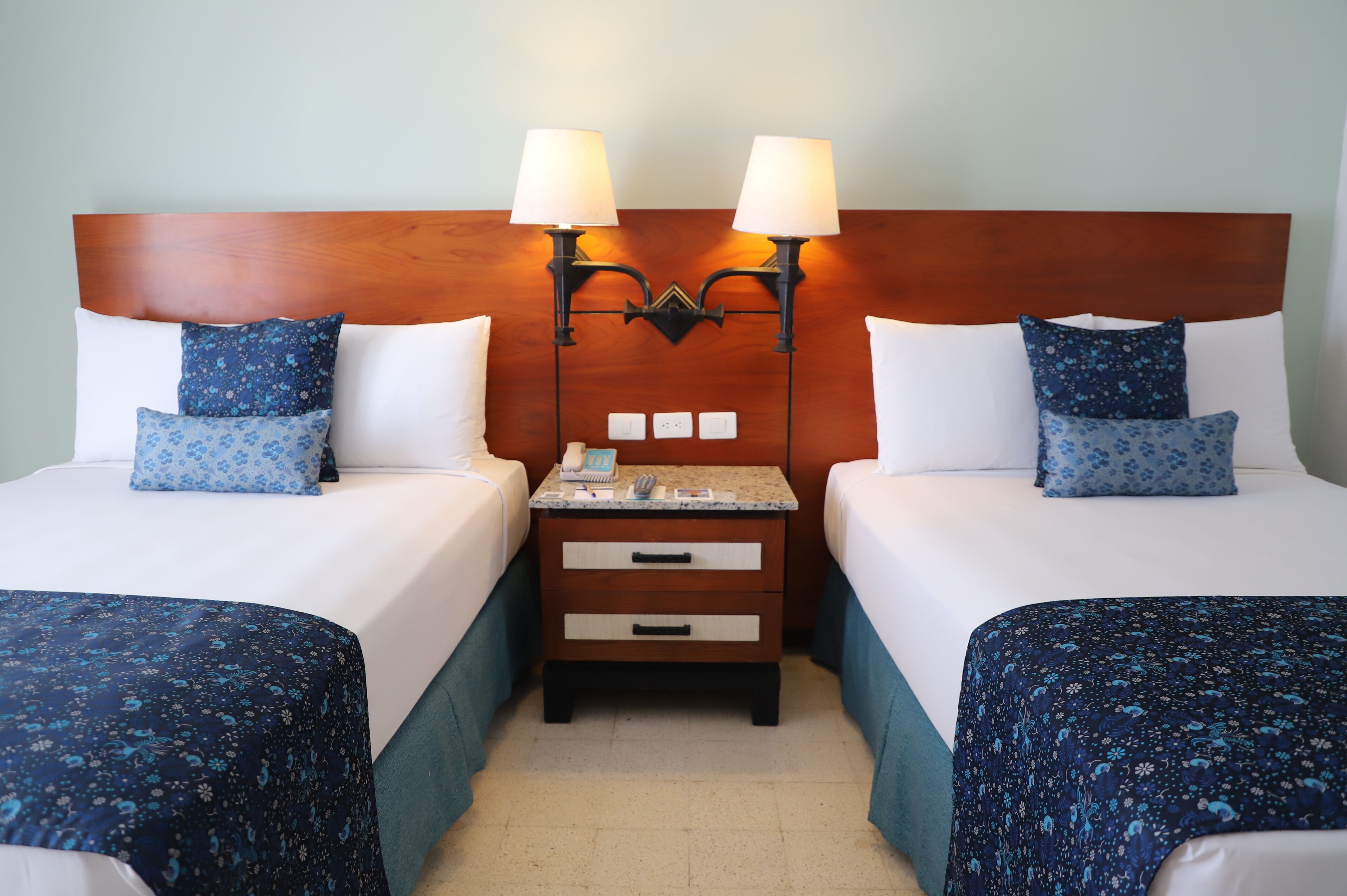 A hotel room with two beds, each with a wooden headboard, white bedding, and blue floral-patterned bedspreads. There are two bedside lamps, a nightstand with drawers, and a small table with a telephone and a lamp.