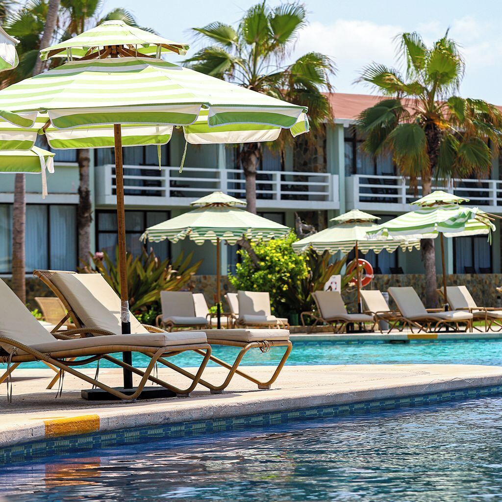 A poolside area with lounge chairs and striped umbrellas, palm trees, and a building in the background.