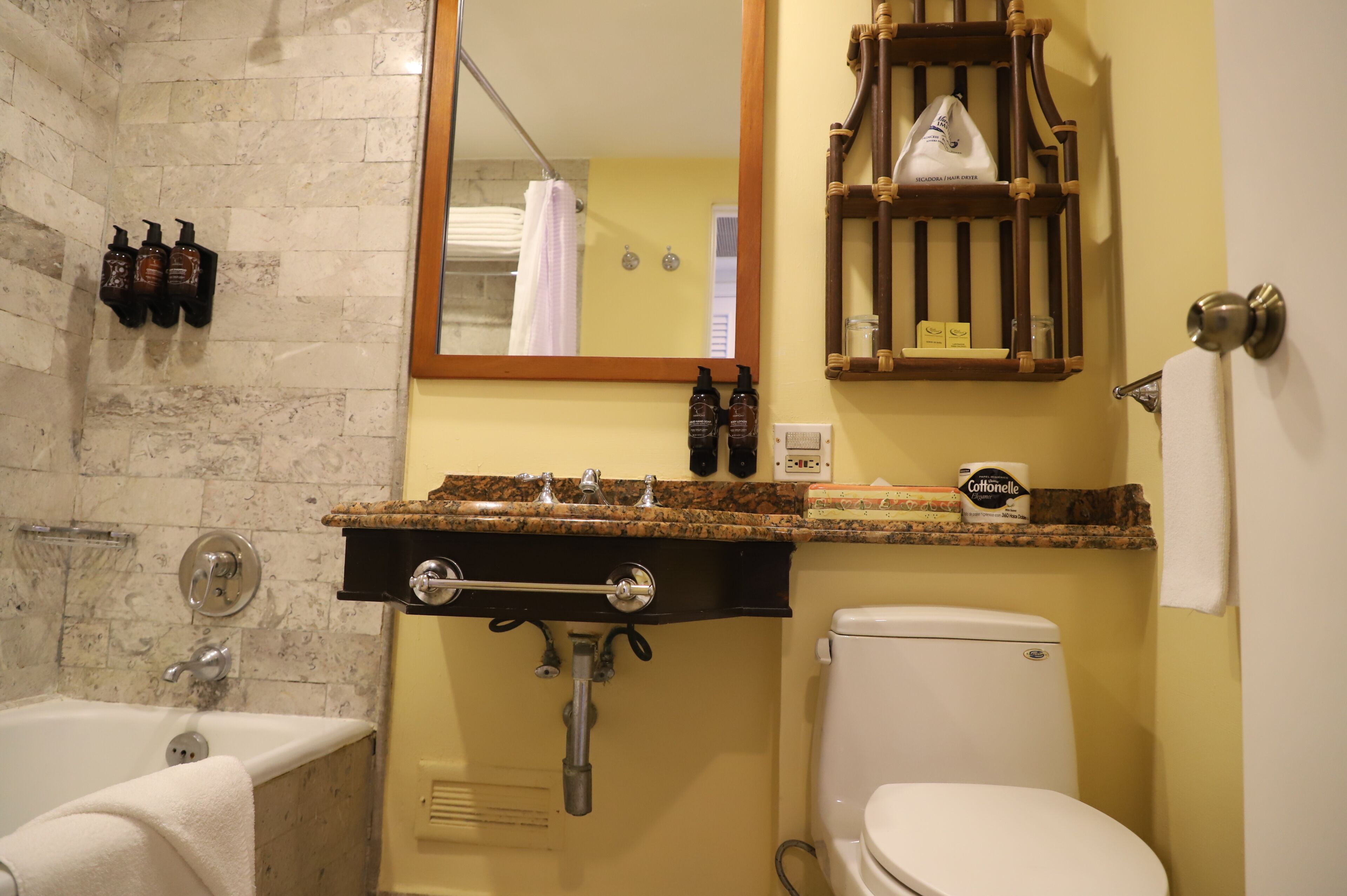 Ein Badezimmer mit Marmorwand, einer Badewanne, einer Toilette, einem Waschbecken und einem Holzregal darüber sowie einem Spiegel.