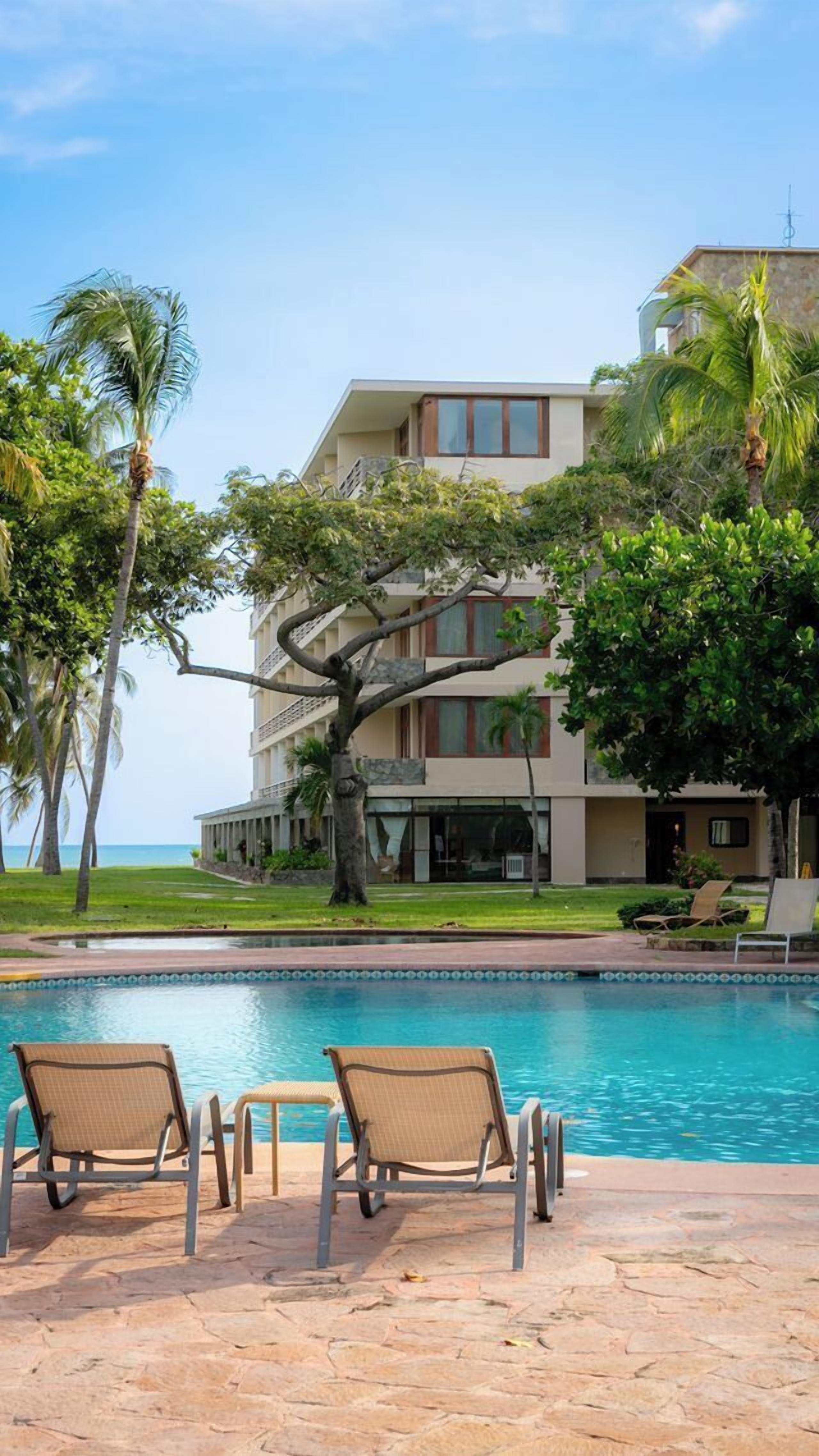 A poolside area with lounge chairs, a building with multiple balconies, and a view of the ocean.