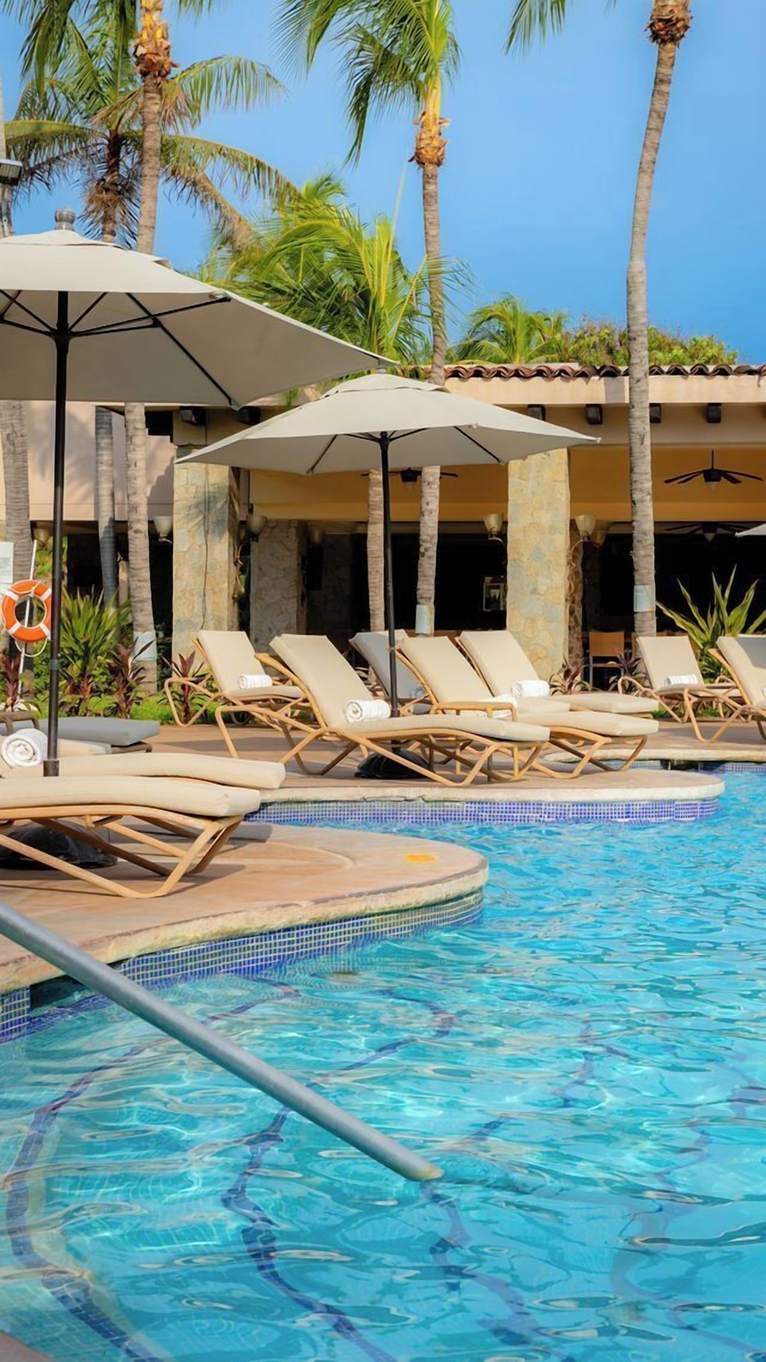 Un área de piscina con sillones reclinables y sombrillas, rodeada de palmeras.