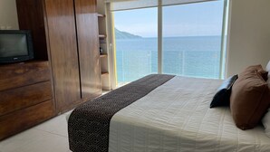 Quarto com uma cama grande, guarda-roupa de madeira, televisão e vista para o mar.