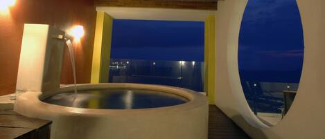 A hot tub with a view of the city at night, a wooden ceiling, and a large arched window.