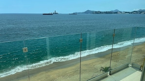 Varanda com vista para a praia e oceano, com guarda-corpo de vidro e céu claro.