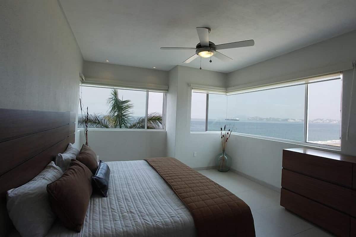 A bedroom with a bed, a dresser, a ceiling fan, and large windows with a view of the sea.