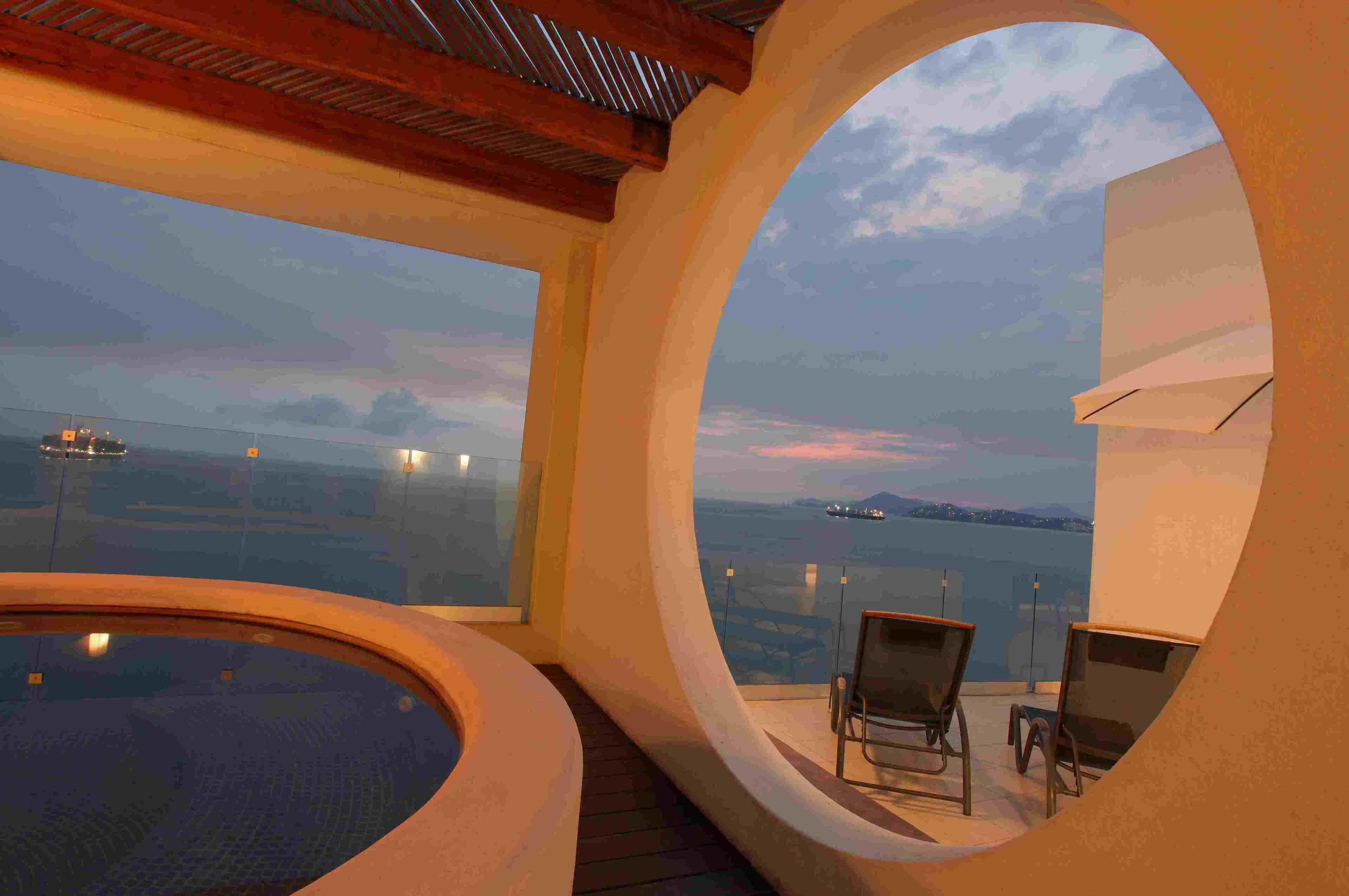A circular room with a hot tub and two chairs facing a large window overlooking a body of water and distant land.